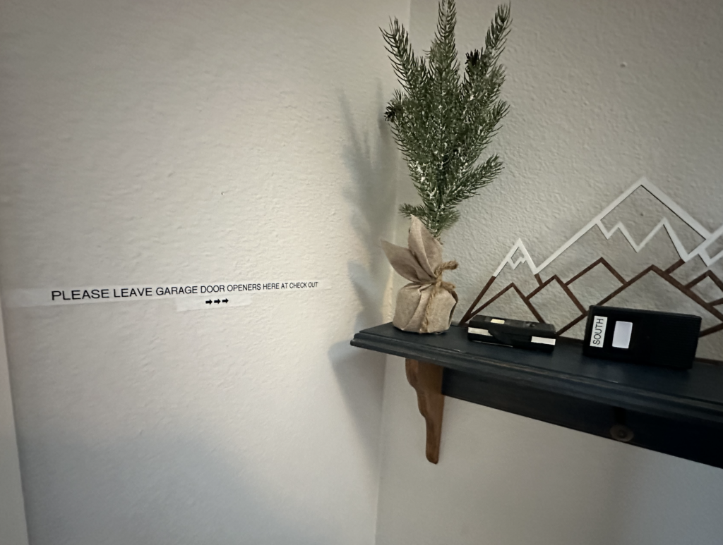 A small shelf with a decorative pine tree in a burlap sack and a garage door opener. Next to it, there is a sign on the wall that reads, 